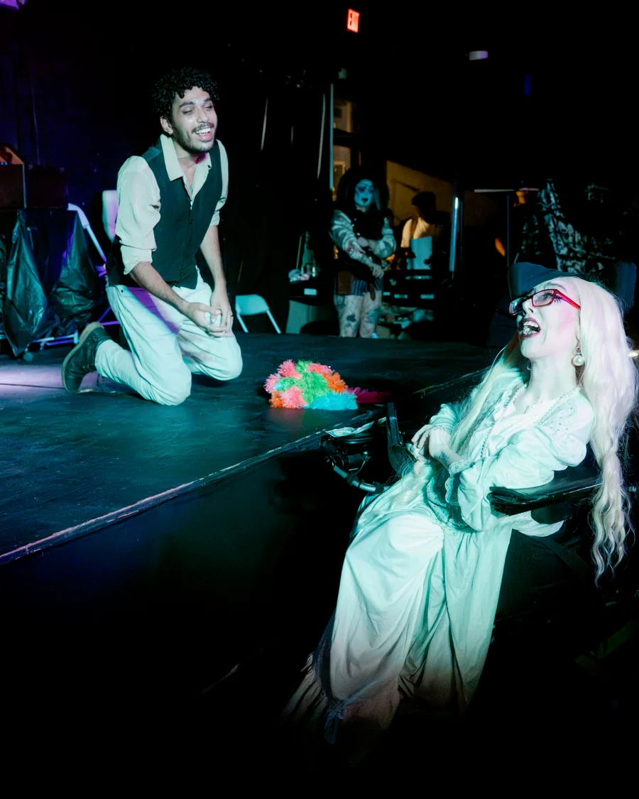 Two Queen-y Todd performers share a scene, one kneeling on stage and one seated beside it.