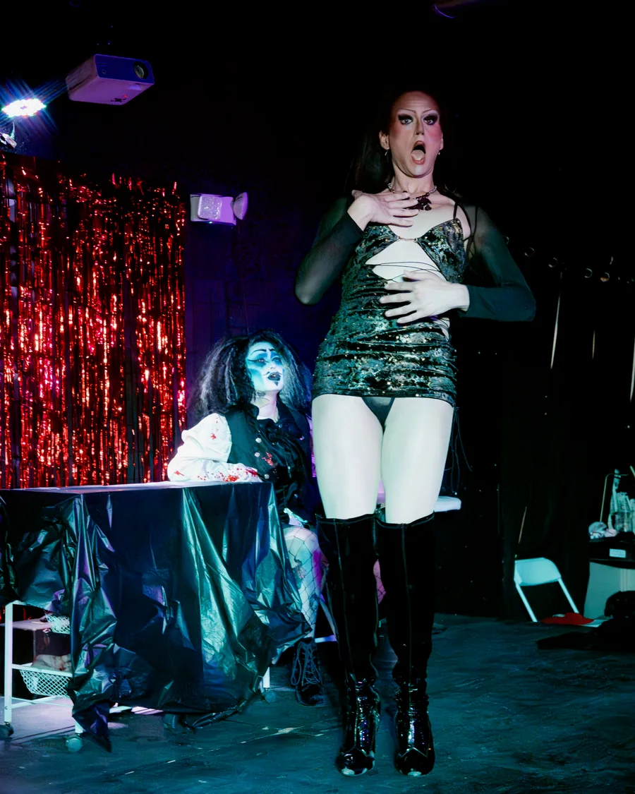 A Queen-y Todd performer in a shimmering costume and tall boots, with a castmate seated behind them.